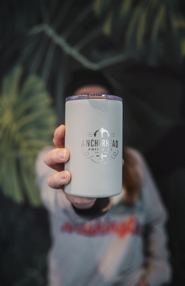 A person holds a gray travel mug with "Anchorhead Coffee Co." logo in focus. The background features blurred tropical leaves, creating a calm, inviting tone.