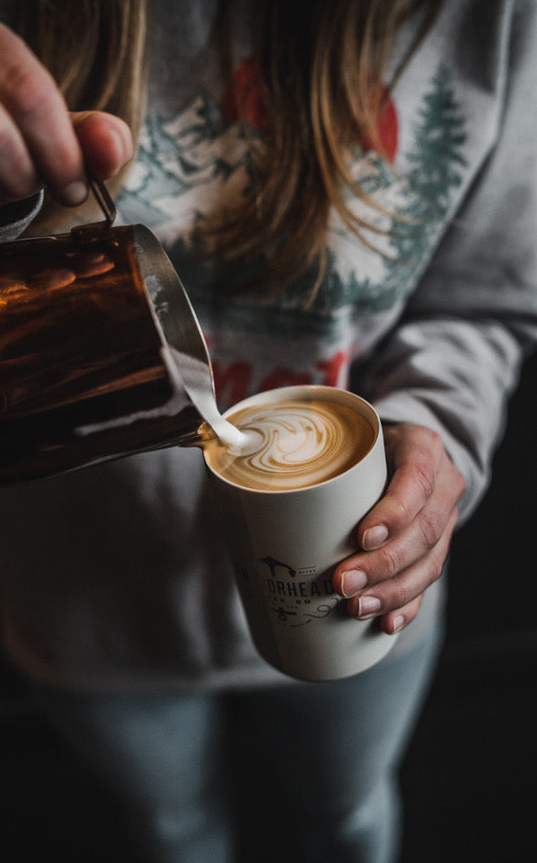 A person wearing a patterned sweater carefully pours steamed milk into a cup of coffee, creating intricate latte art. The scene feels cozy and artisanal.