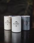 Three ceramic travel mugs with the Atlantis brand logo, in white, black, and silver colors, displayed on a dark background.